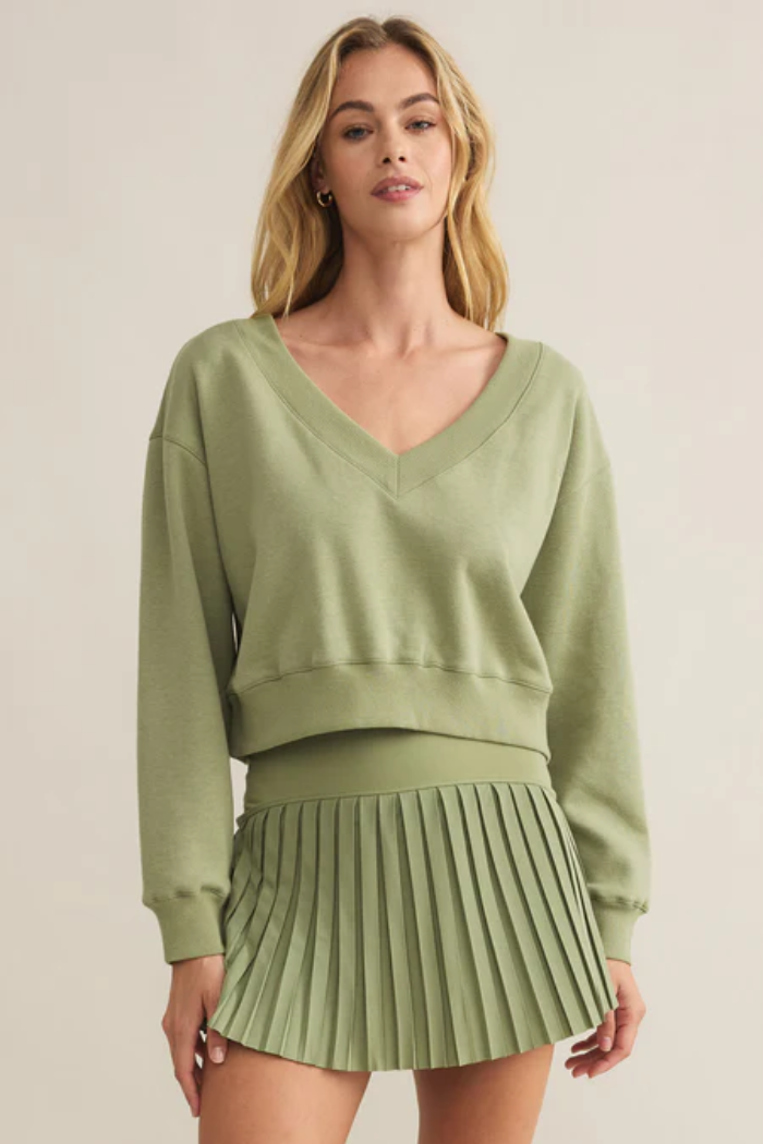 A woman wearing a green v-neck, long-sleeved cropped sweatshirt with a pleated green mini skirt.