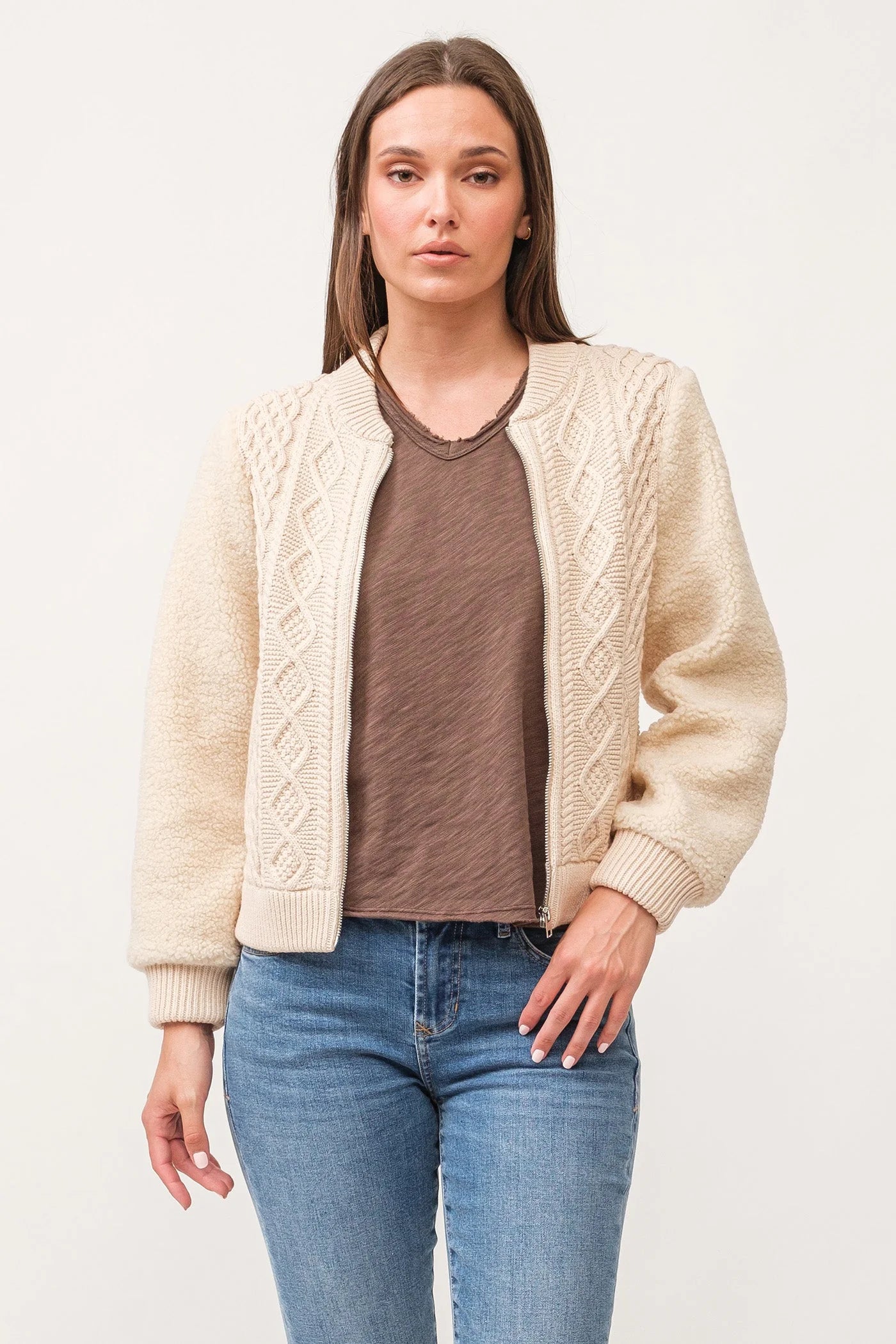 Woman wearing a cream cable knit cardigan over a brown shirt and blue jeans on a white background