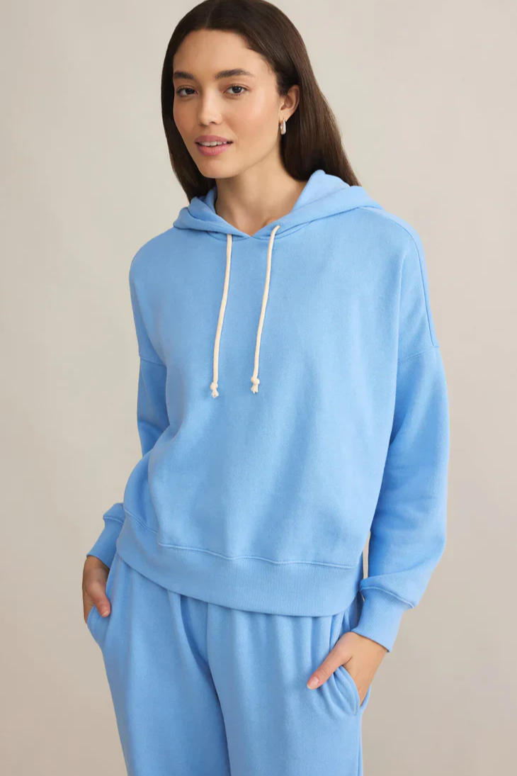 A woman wearing a blue hooded hoodie with the drawstrings down, paired with blue sweatpants, standing against a neutral background.