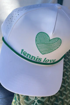 White cap with green heart design and 'tennis love' text on a blurred background