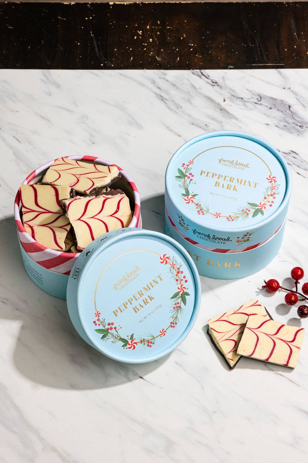 Two open light blue tins of French Broad peppermint bark on white marble with holiday berries