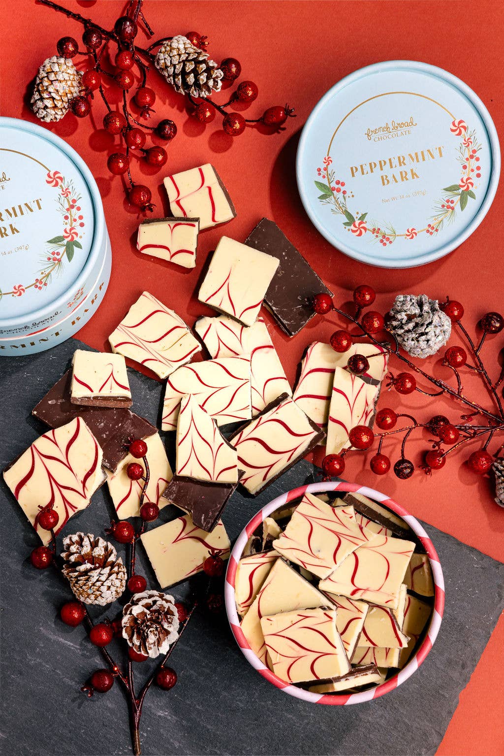 Scattered white and dark chocolate peppermint bark pieces with red swirls, pine cones, and holiday tins