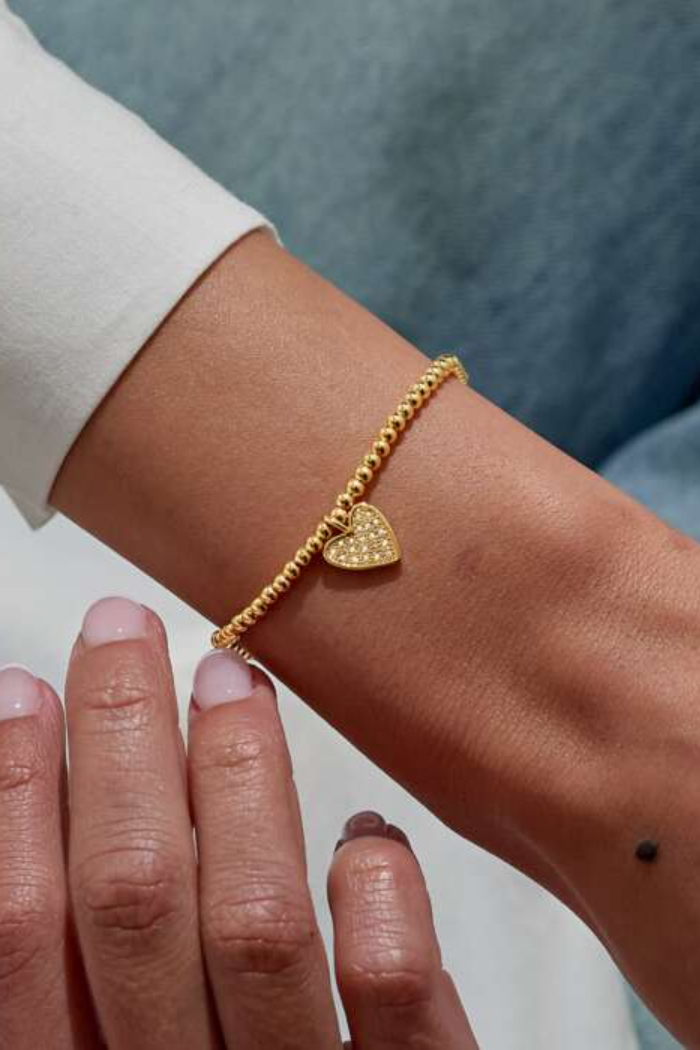 Gold bracelet with a heart charm on a wrist against a neutral background
