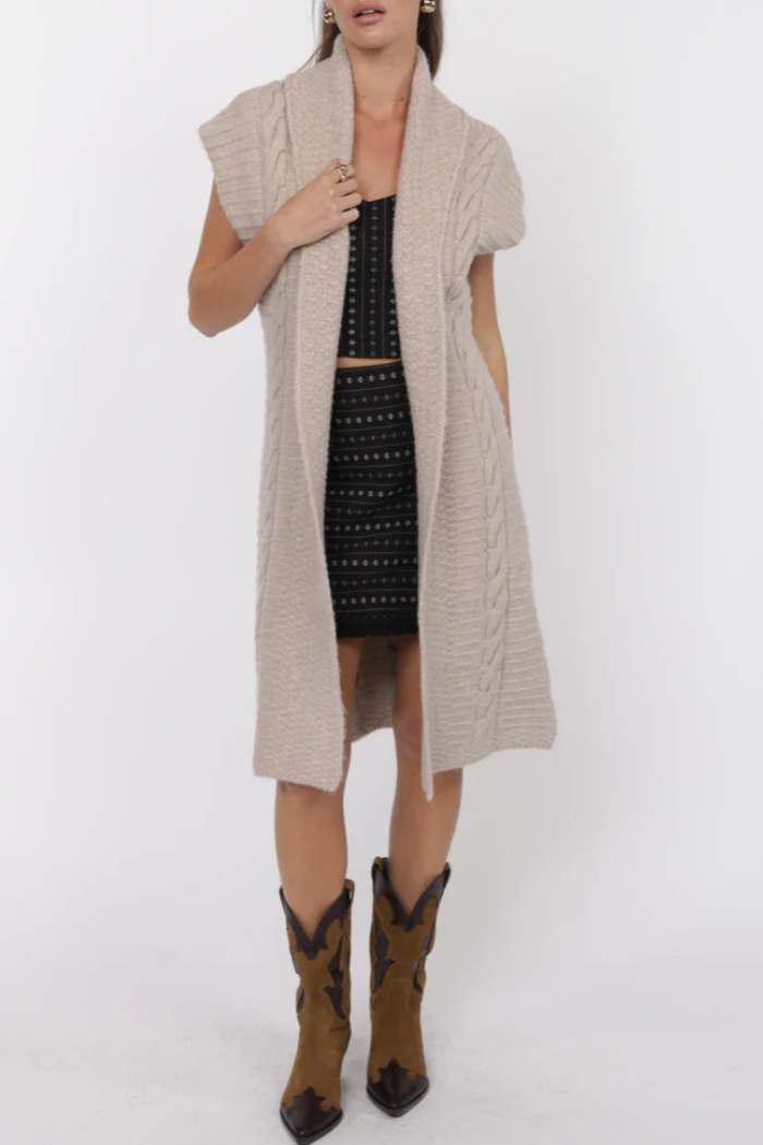 Woman wearing a beige long vest over a black patterned dress with brown cowboy boots on a white background