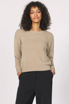 Woman wearing a beige sweater and black pants against a white background