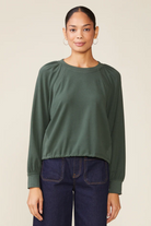 Woman wearing a green long-sleeve top and dark jeans against a beige background